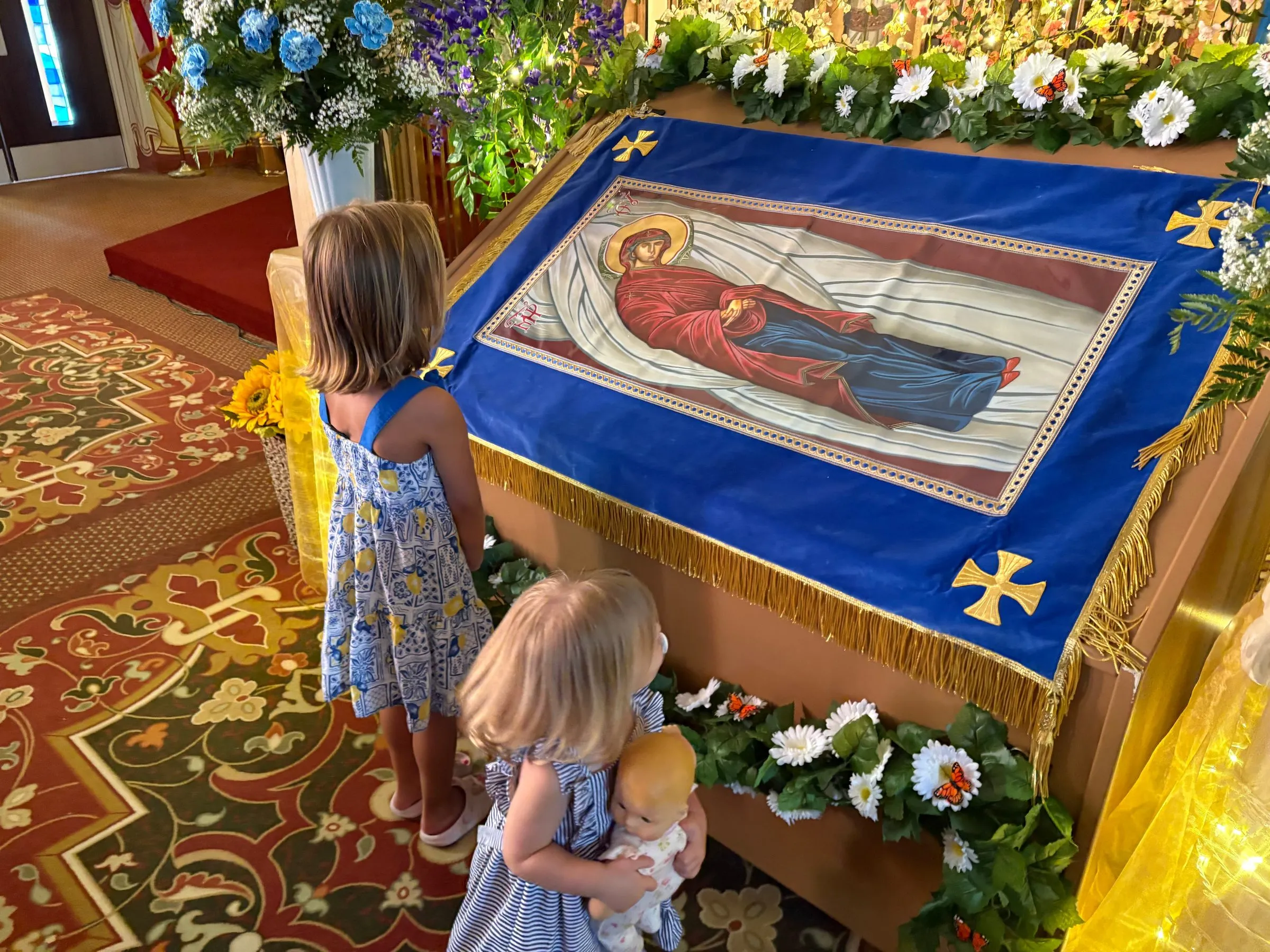 Two small girls standing before the flower-decked icon of the Dormition of the Mother of God